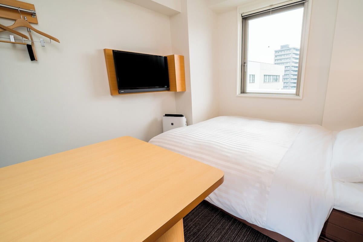 A hotel room with a bed, a wooden table, a TV mounted on the wall, and a window with a view of buildings.