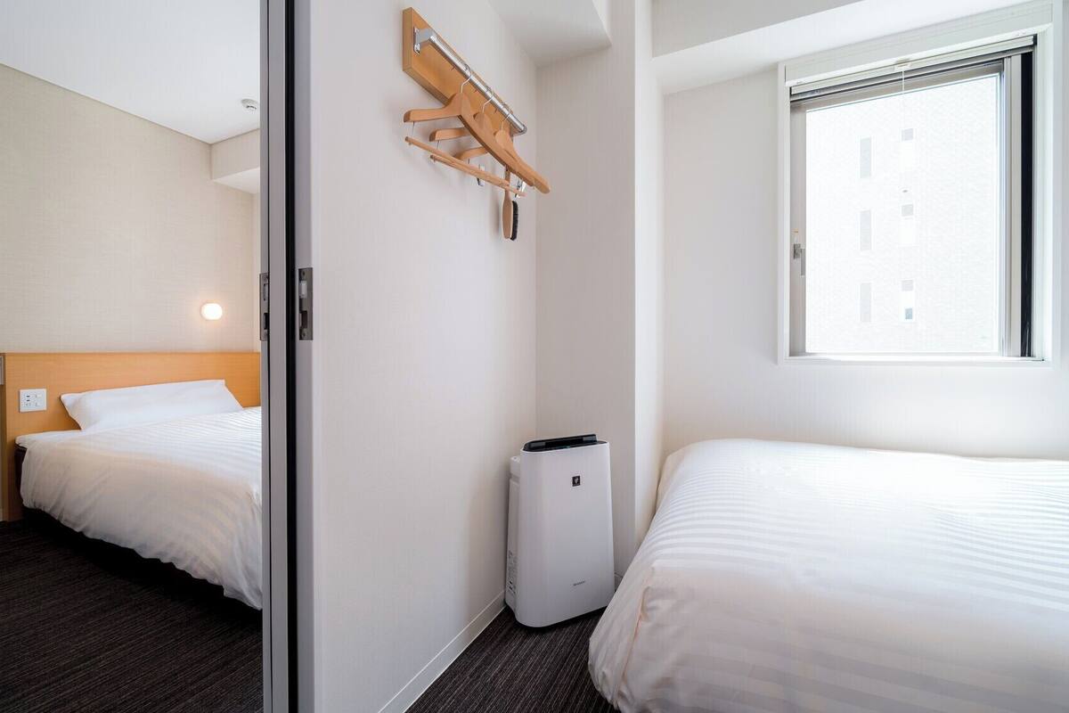 A hotel room with a bed, a desk, a chair, a coat rack, a window, and a wall-mounted heater.