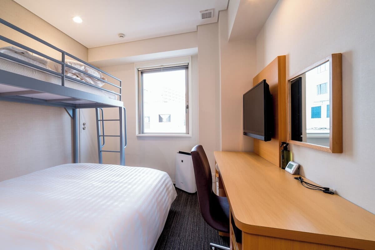 A hotel room with bunk beds, a desk, a chair, a television, and a window.