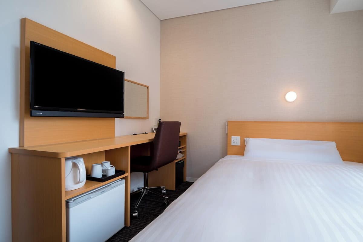A hotel room with a bed, a desk with a flat-screen TV, a chair, a mini-fridge, and a wall-mounted light.