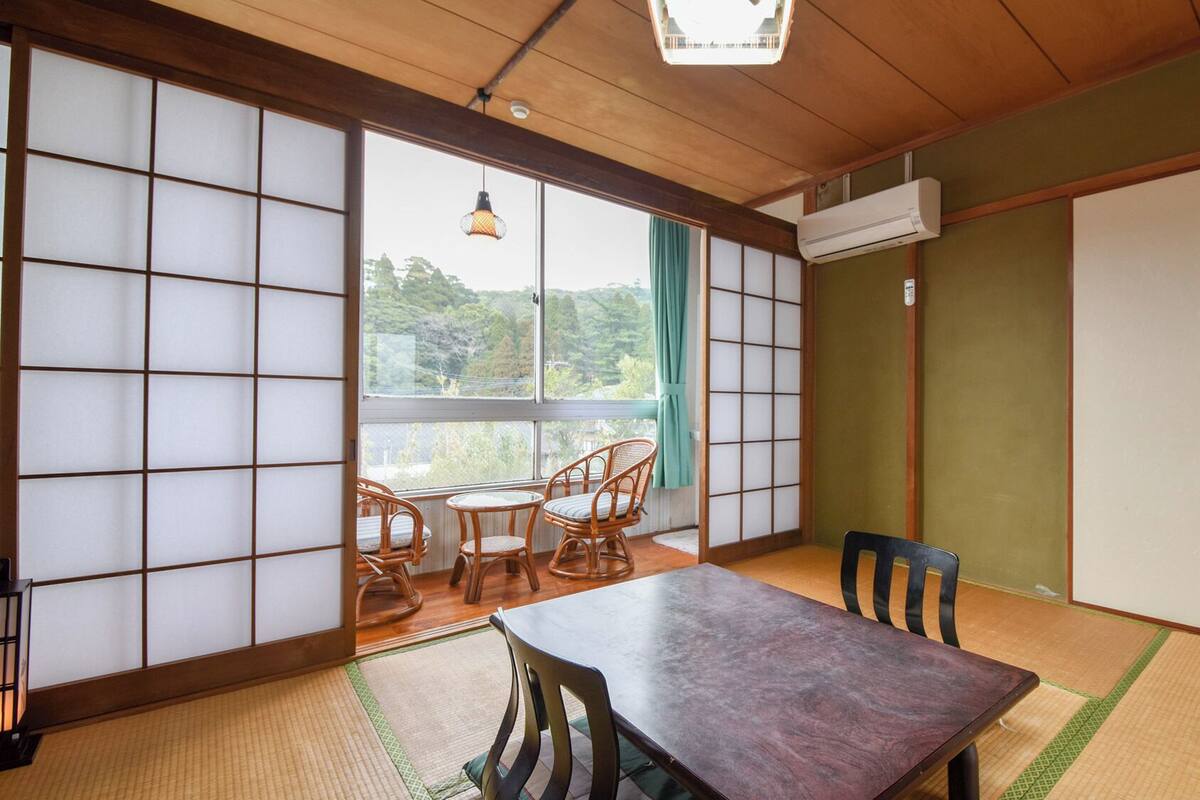 Una habitación japonesa tradicional con suelo de tatami, puertas correderas, una mesa con sillas y vistas a la vegetación exterior.