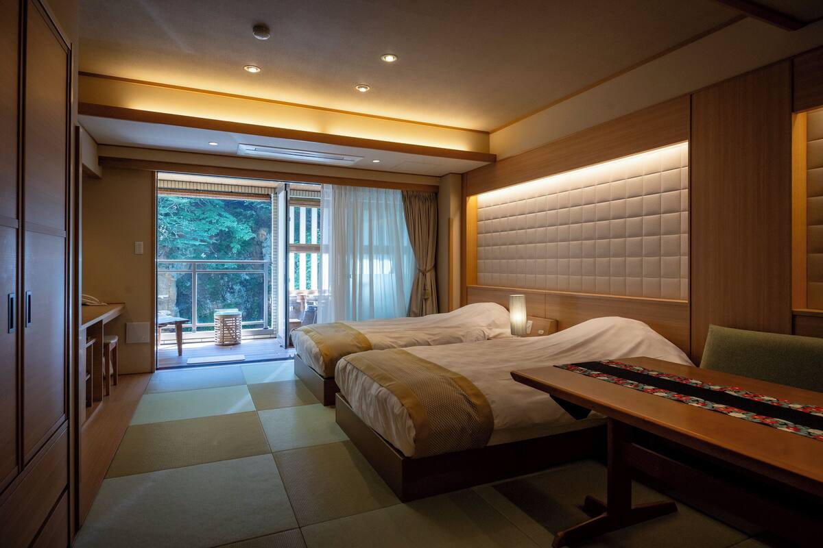 A hotel room with two beds, a wooden desk, a large window with curtains, and a view of greenery outside.