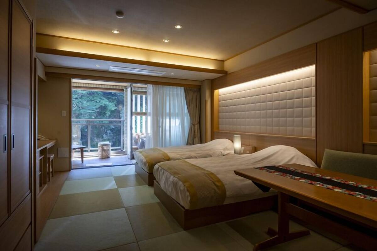 A hotel room with two beds, a wooden desk, a large window with curtains, and a view of greenery outside.
