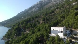 A white building with a flat roof, surrounded by dense green trees and rocky mountains. - Luxury apartment INA (Omis)