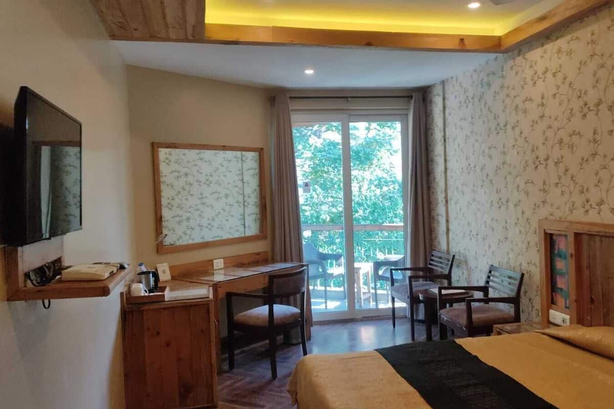 A hotel room with a bed, desk, chairs, a television, and a balcony with a view of trees.