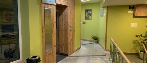 Un pasillo con paneles de madera, un cubo de basura, una planta en maceta y una escalera con barandales verdes.