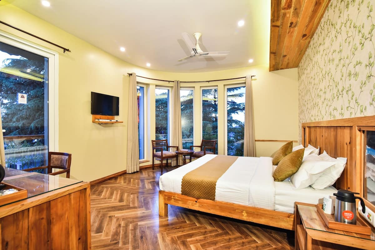 A bedroom with a wooden bed, a TV, a desk, and large windows with a view of trees.
