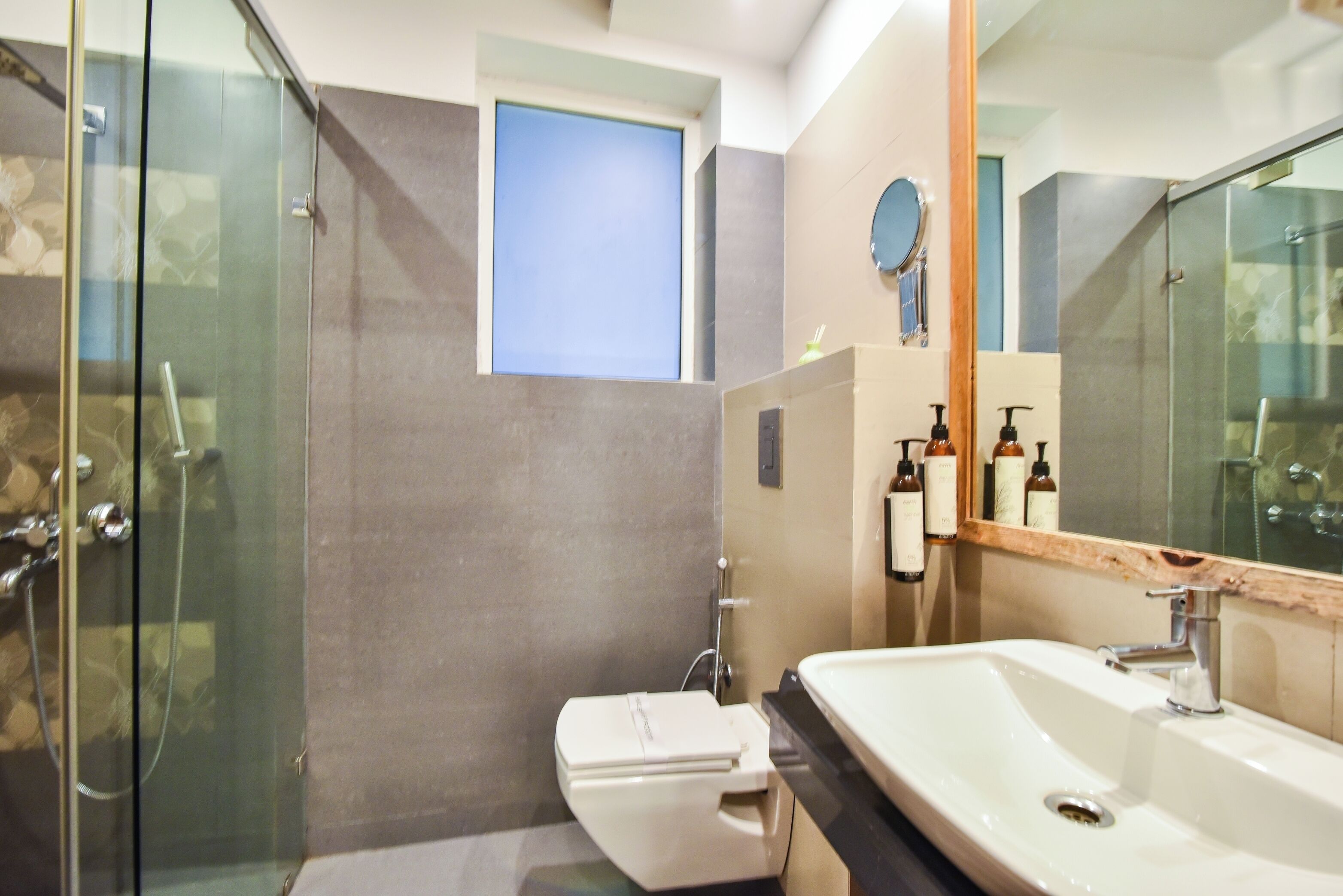 A modern bathroom with a glass shower enclosure, a toilet, a sink with a mirror, and a small window.