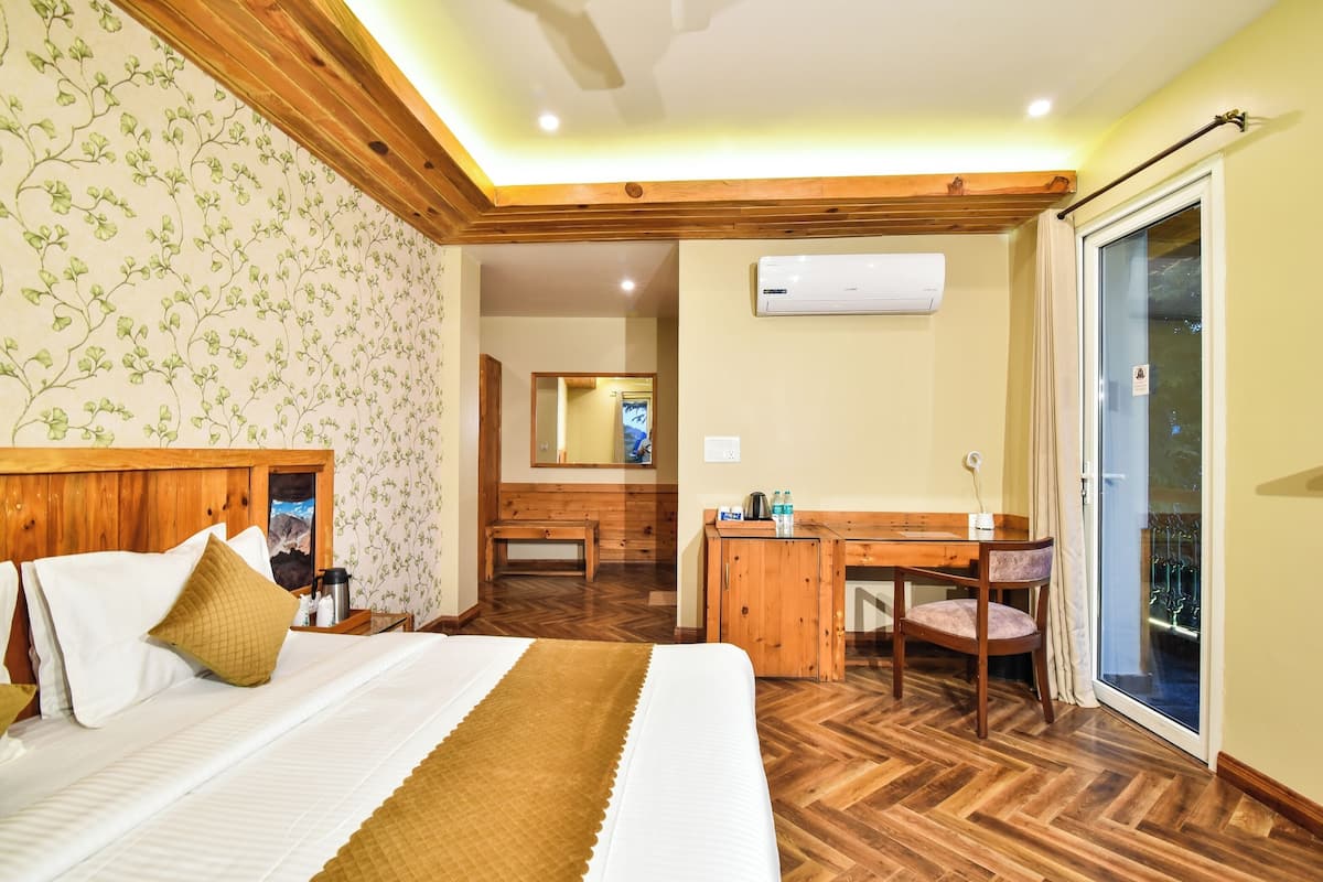 A hotel room with a bed, wooden headboard, a desk with a chair, a small table with a chair, a mirror, and a sliding glass door leading to an outdoor area.