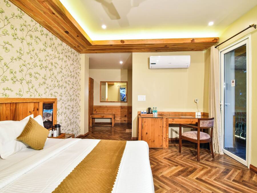 A hotel room with a bed, wooden headboard, a desk with a chair, a small table with a chair, a mirror, and a sliding glass door leading to an outdoor area.