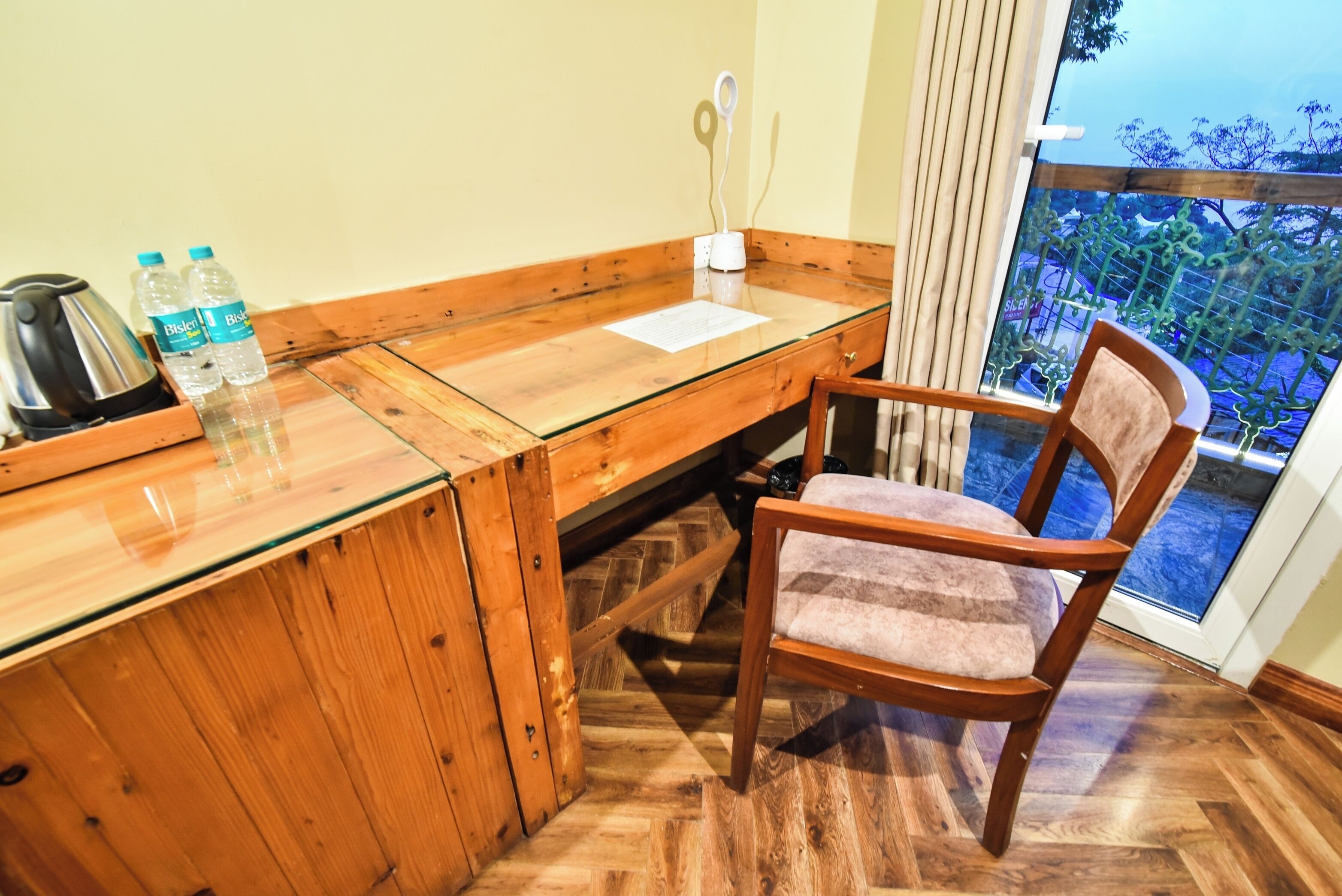 A wooden desk with a glass top, two water bottles, a kettle, and a chair with a cushioned seat.