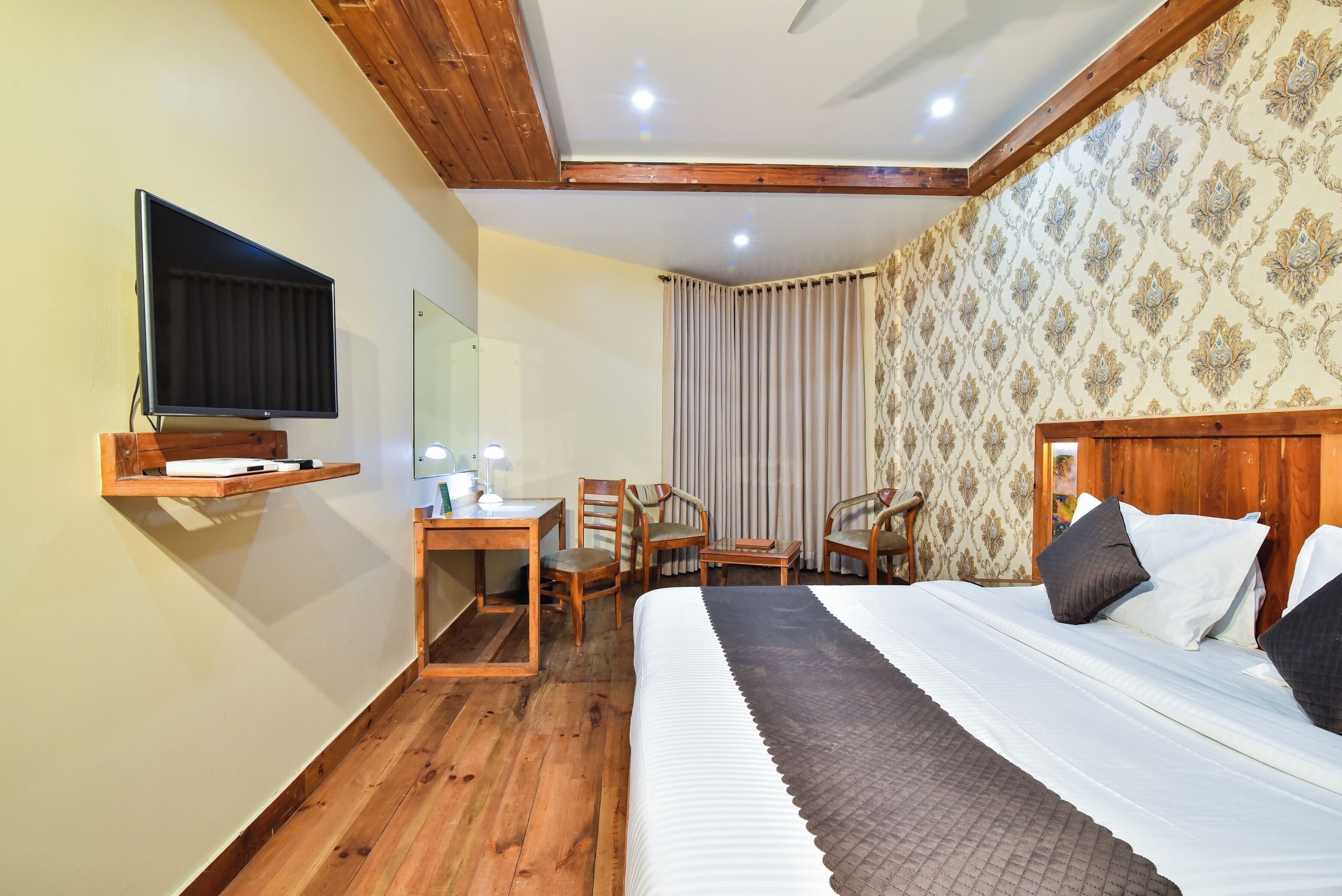 A hotel room with a wooden ceiling, a bed with white bedding, a desk with a lamp, two chairs, and a TV mounted on the wall.