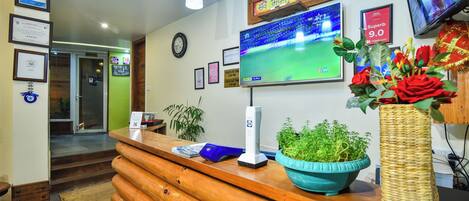 Un espace d’accueil avec un comptoir en bois, un téléviseur diffusant un match de cricket, des certificats encadrés et une horloge au mur.