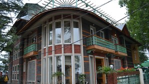 A two-story building with a curved facade, large windows, and a balcony with green railings. - D'S CASA Hotel (Dharamshala)