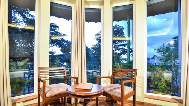 A room with a round wooden table and two chairs, large windows with a view of trees and a garden. - D'S CASA Hotel (Dharamshala)