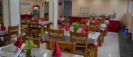 Un comedor con varias mesas, cada una con sillas de madera, manteles rojos y servilletas rosas.