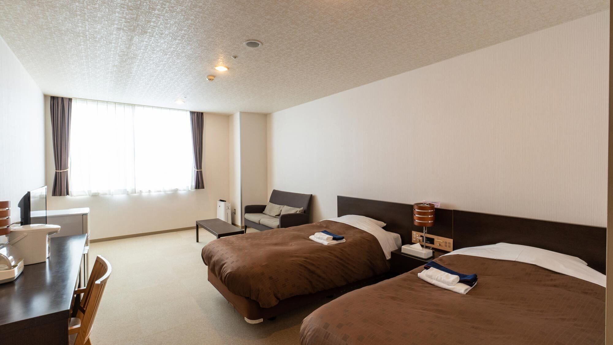Habitación de hotel con dos camas, un escritorio, una silla y un ventanal con cortinas.