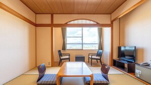 Una habitación japonesa tradicional con suelo de tatami, una mesa de madera baja, dos sillas, un televisor y vistas a las montañas a través de un ventanal grande.