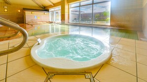 Un jacuzzi en un amplio espacio interior con piscina, grandes ventanales y suelo de baldosas.