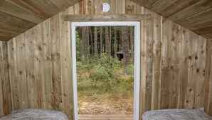 Een houten hutje met twee bedden, een deur die uitkomt op een bebost gebied en een kleine schuur op de achtergrond.