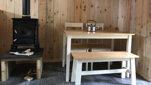Een houten hut van binnen, met een zwart houten kachel, een witte tafel met banken, en een waterkoker op de tafel.