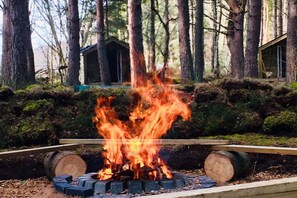 A campfire surrounded by logs and a forested area with a small cabin in the background.