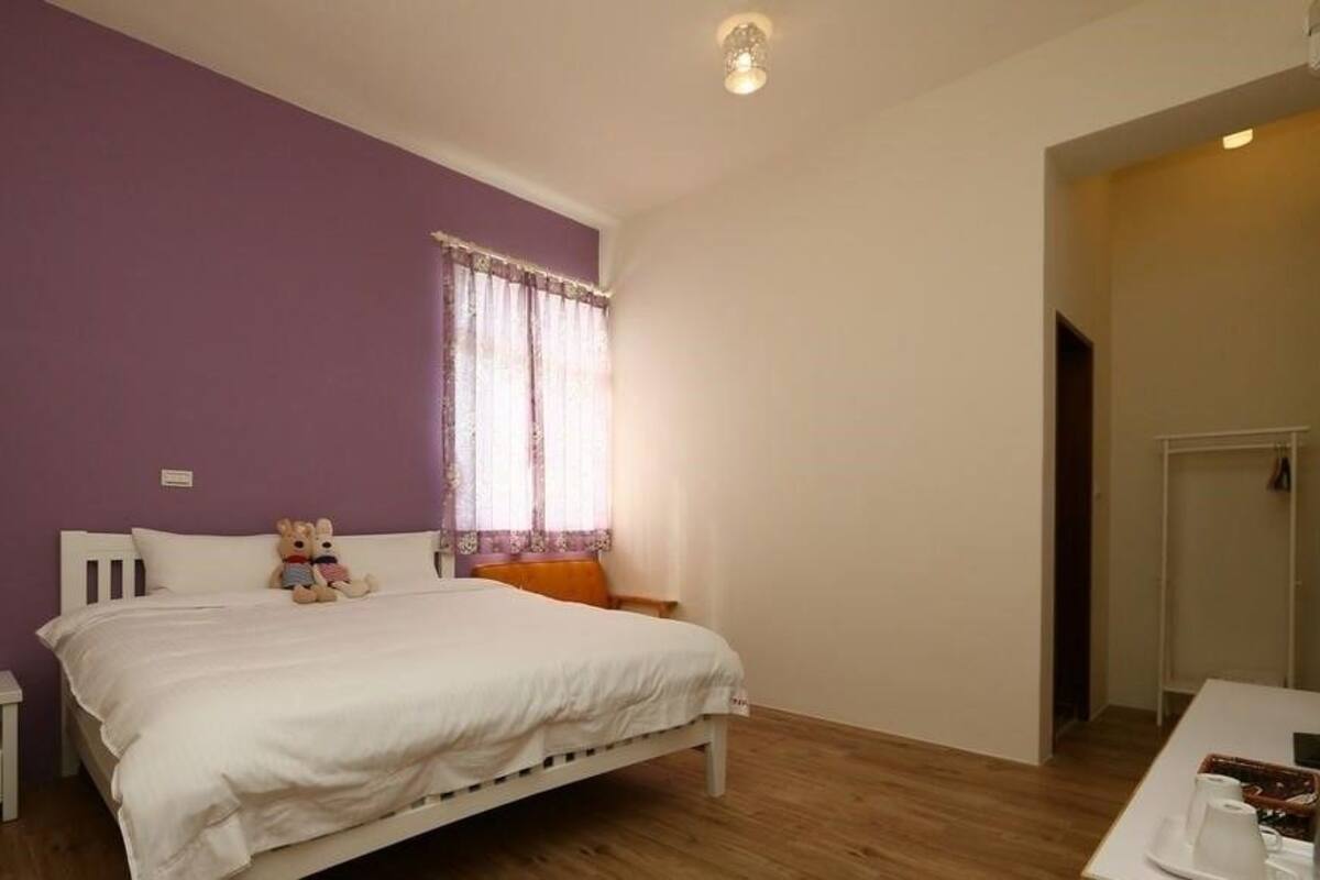 A bedroom with a bed, a nightstand, a window with curtains, and a small table with a cup and saucer.
