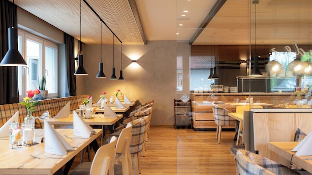 A modern restaurant with wooden tables and chairs, pendant lighting, and a view of the kitchen. - Hotel Landgasthof Linde (Deggenhausertal)