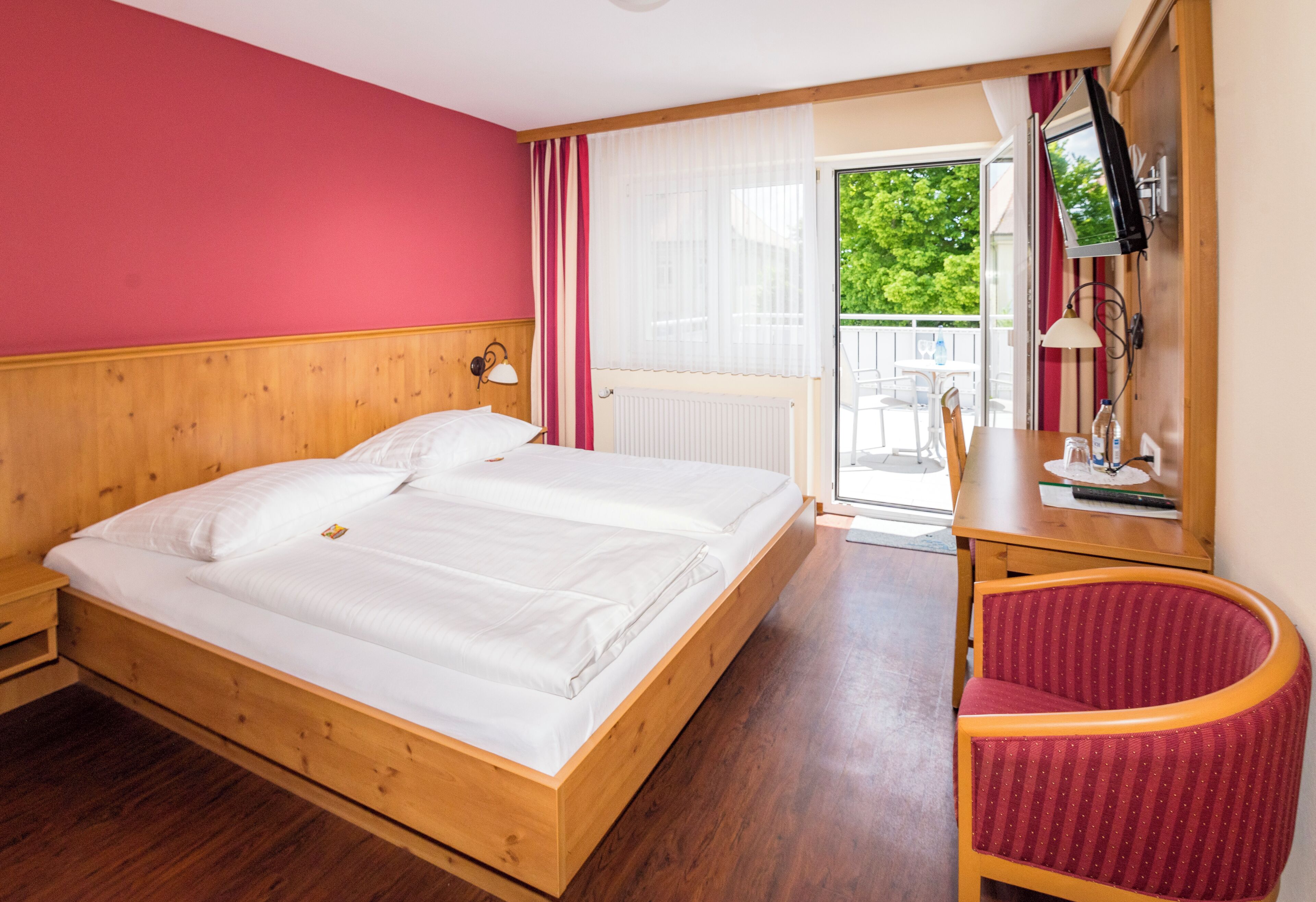 A double bed with wooden headboard, a red chair, a wooden desk with a lamp, and a sliding glass door leading to a balcony.