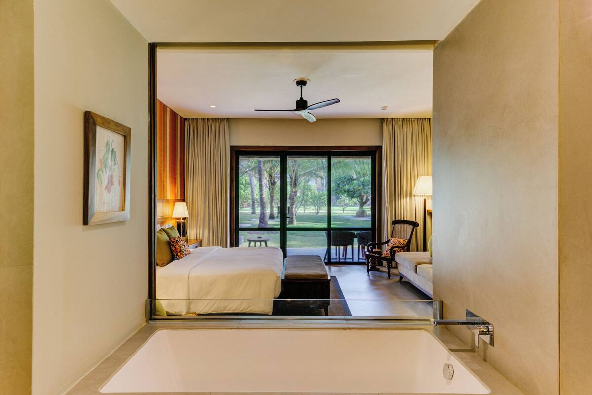 Una habitación de hotel con una cama grande, una zona de estar con una silla y una mesa, una bañera y vistas al exterior a través de amplios ventanales.