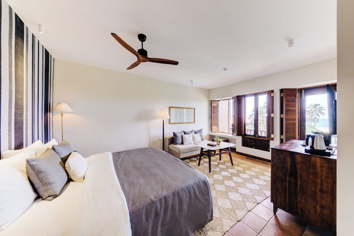 Habitación de hotel con cama, escritorio, silla, ventilador de techo y ventanas grandes con contraventanas.