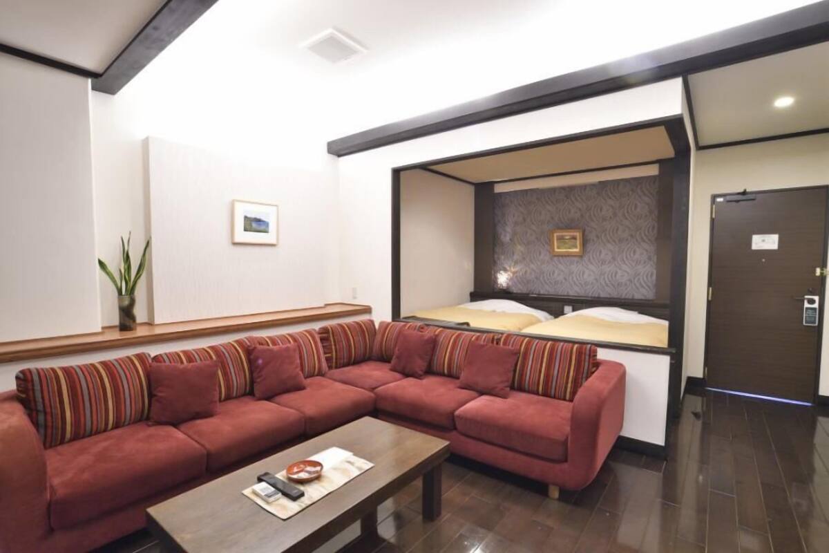 A modern hotel room with a sectional sofa, a small table, a bed with a headboard, and a wall-mounted picture.