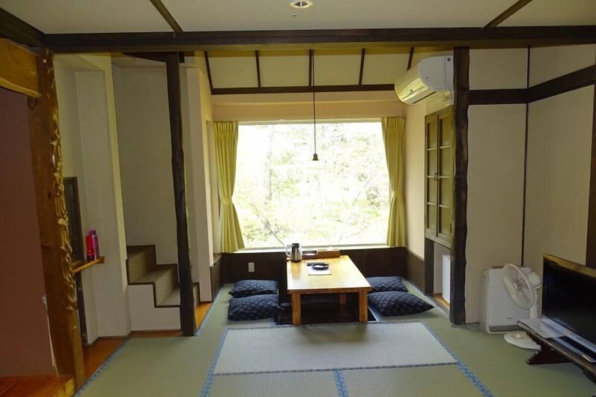 A traditional Japanese-style room with tatami flooring, a low table with cushions, a window with curtains, and a ceiling fan.