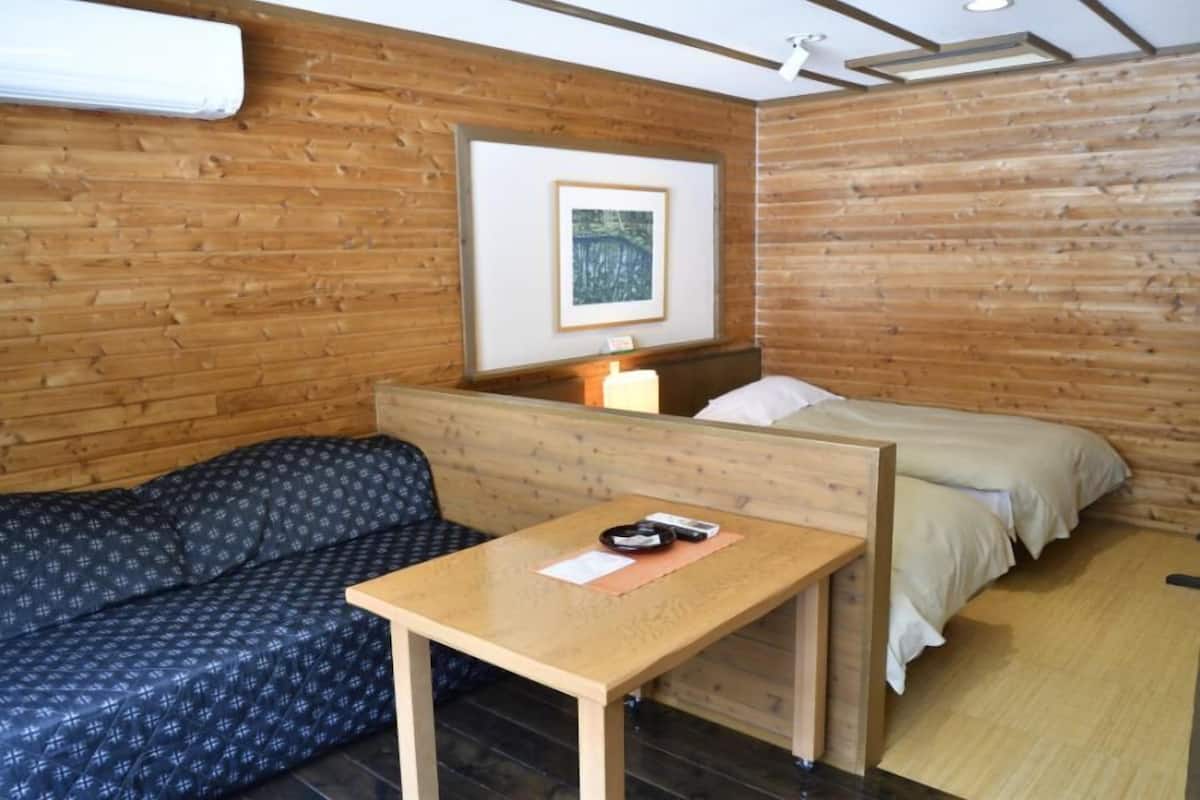 A room with a wooden wall, a bed, a sofa, and a table with a lamp.