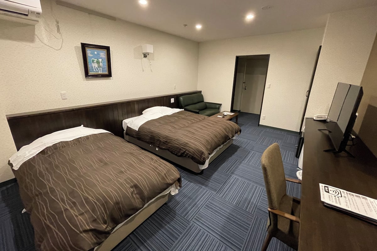 A hotel room with two beds, a desk, a chair, a TV, and an air conditioning unit.