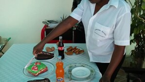 A person serving food on a table with plates of pastries, a bottle of soda, and a glass of water. - Cingaki Hotel (Mombasa)