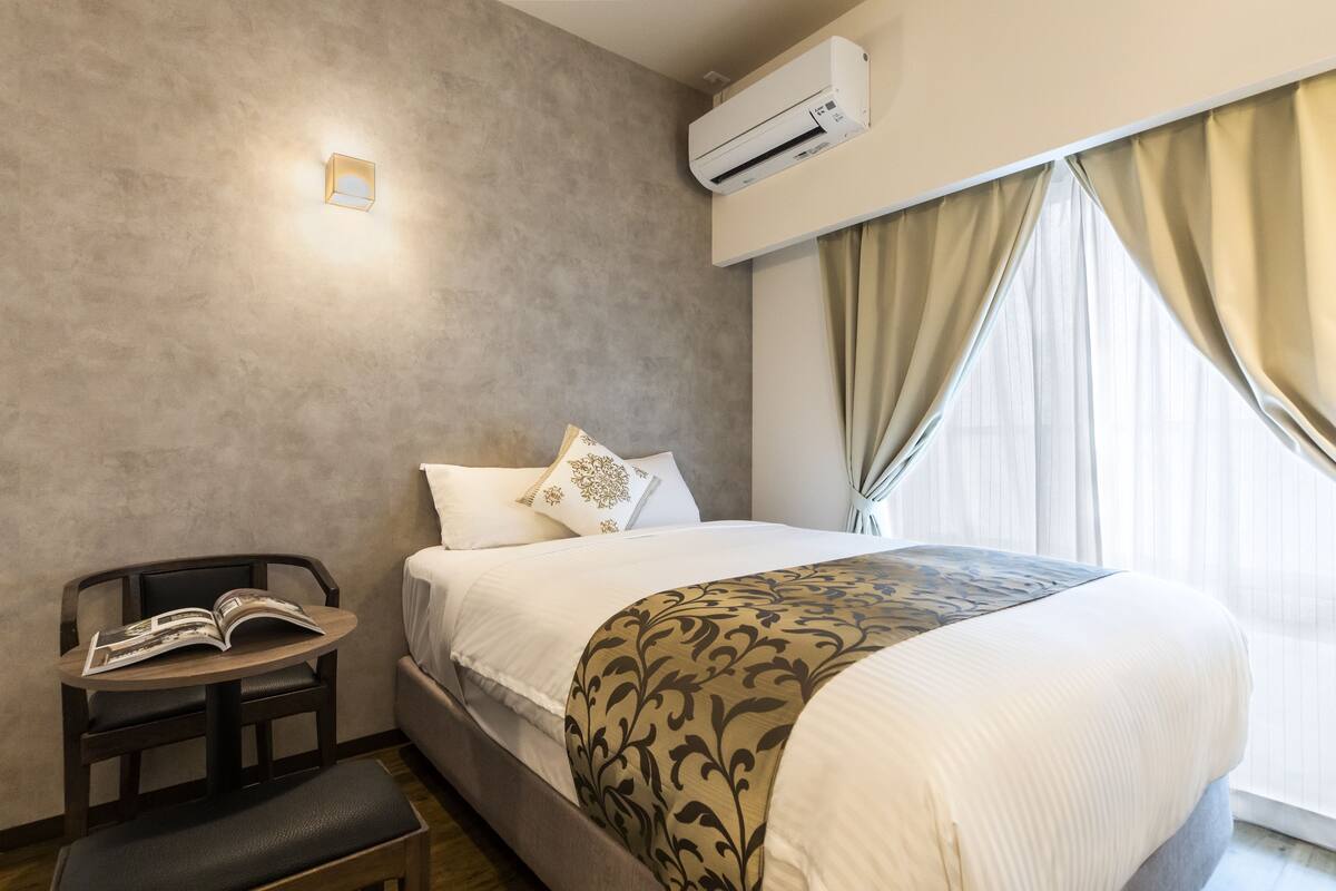 Quarto de hotel com uma cama, mesa de cabeceira com uma revista, uma cadeira e uma janela com cortinas.