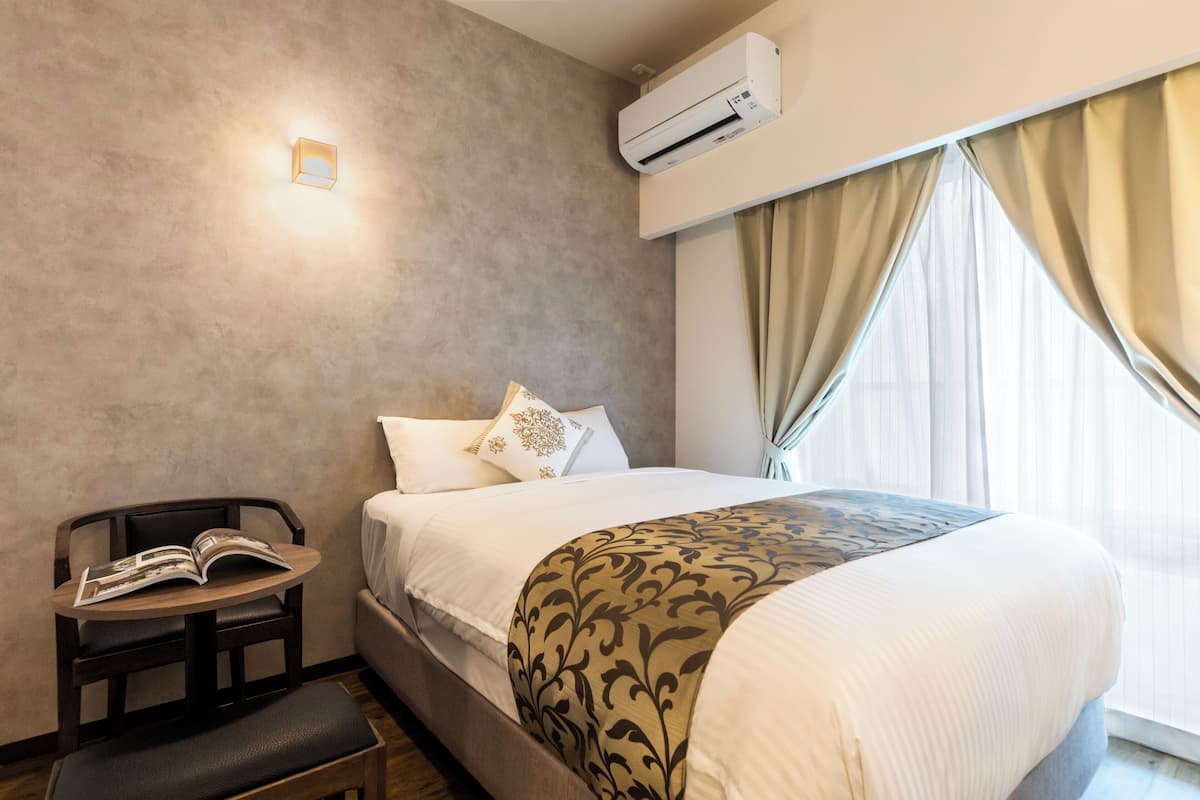 Quarto de hotel com uma cama, mesa de cabeceira com uma revista, uma cadeira e uma janela com cortinas.