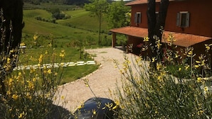 A rural house with a gravel driveway leading to a red-roofed building, surrounded by green fields and yellow flowering plants. - Villa del Sole (San Gimignano)