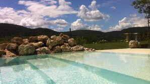A clear pool with a rocky edge, surrounded by a grassy area and hills in the background. - Villa del Sole (San Gimignano)