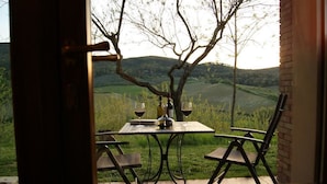 A dining table with four chairs, a bottle, and two wine glasses, set against a backdrop of rolling hills and a tree. - Villa del Sole (San Gimignano)