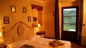 A bedroom with a bed, a nightstand, a lamp, a small table with a vase, and a window with a view of greenery. - Villa del Sole (San Gimignano)