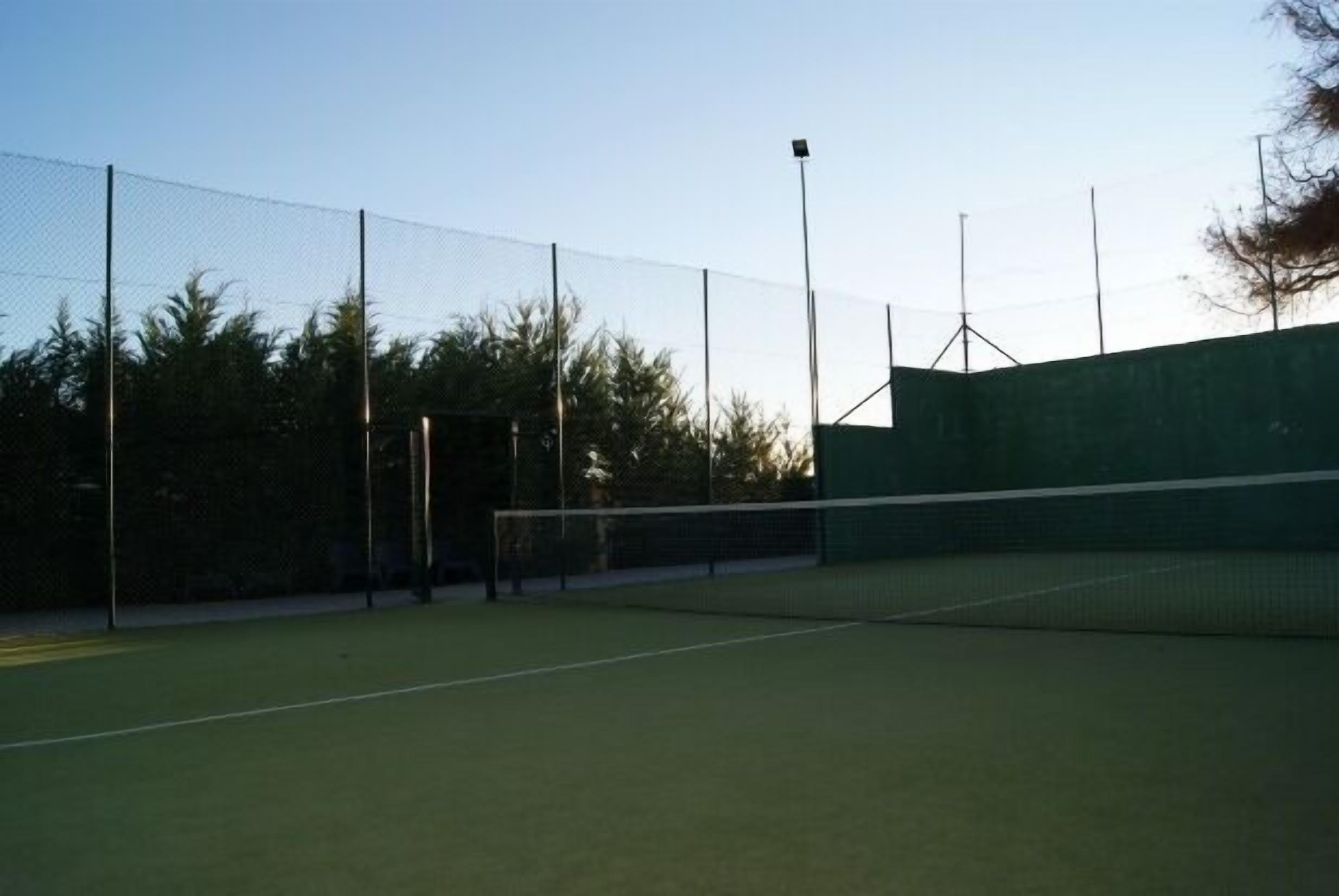 An outdoor tennis court with a net, surrounded by a chain-link fence and trees.