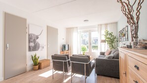 A modern living room with a sofa, two armchairs, a coffee table, and a TV. There is a window with curtains, a plant, and a decorative vase on a wooden cabinet. - Holiday home for 6 people in Wemeldinge (Wemeldinge)