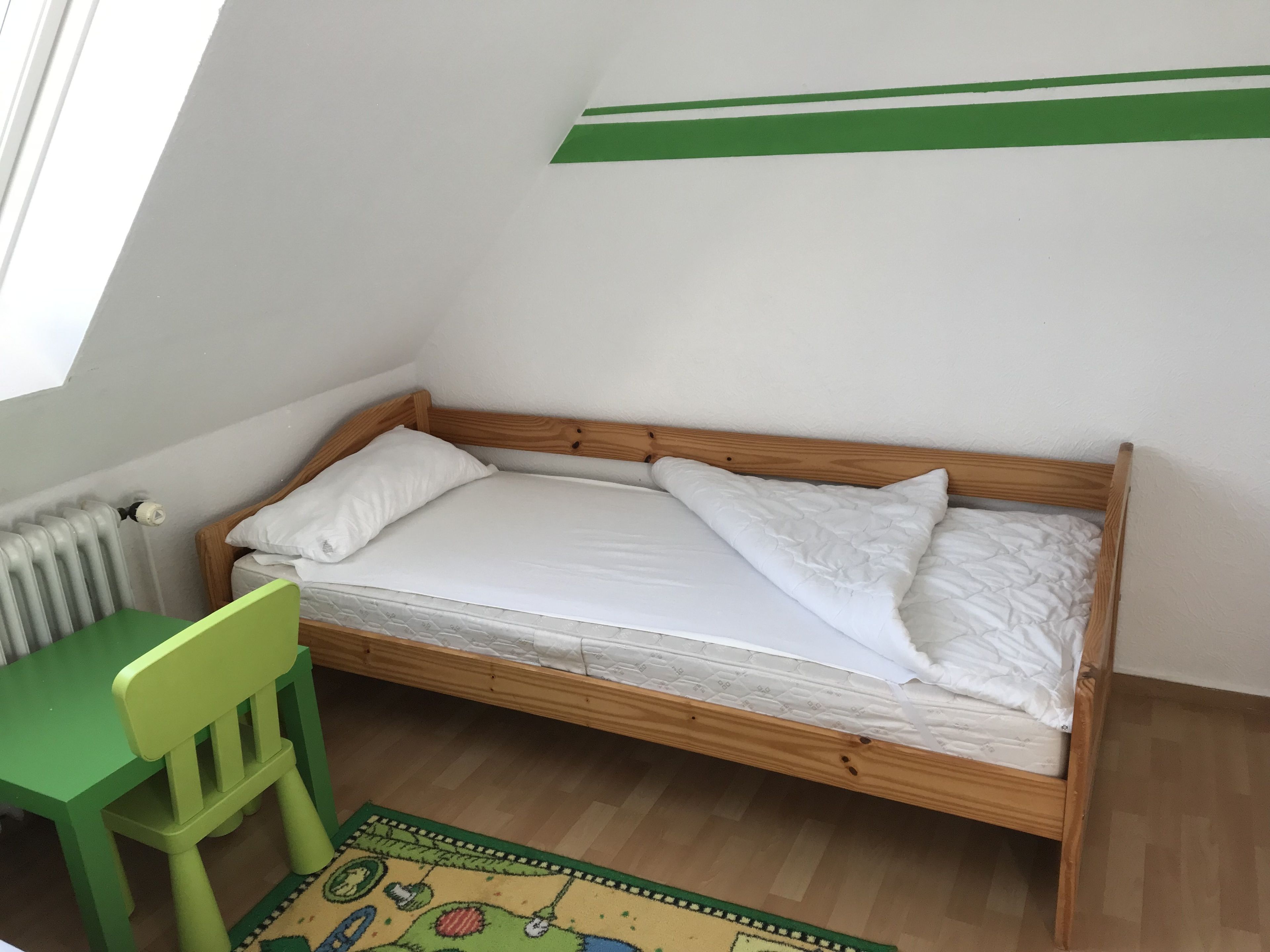 A small, single-bed room with a wooden frame, a green chair, and a radiator.