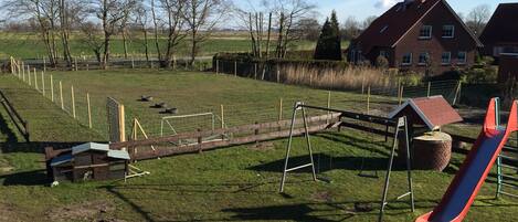 A playground with a slide and swings, surrounded by a fenced grassy area and trees.
