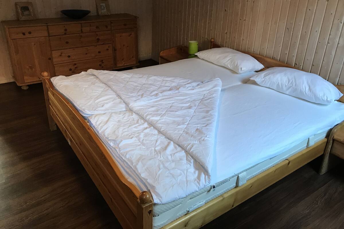Puinen sänky valkoisilla vuodevaatteilla huoneessa, jossa on puuseinät ja puinen lipasto.