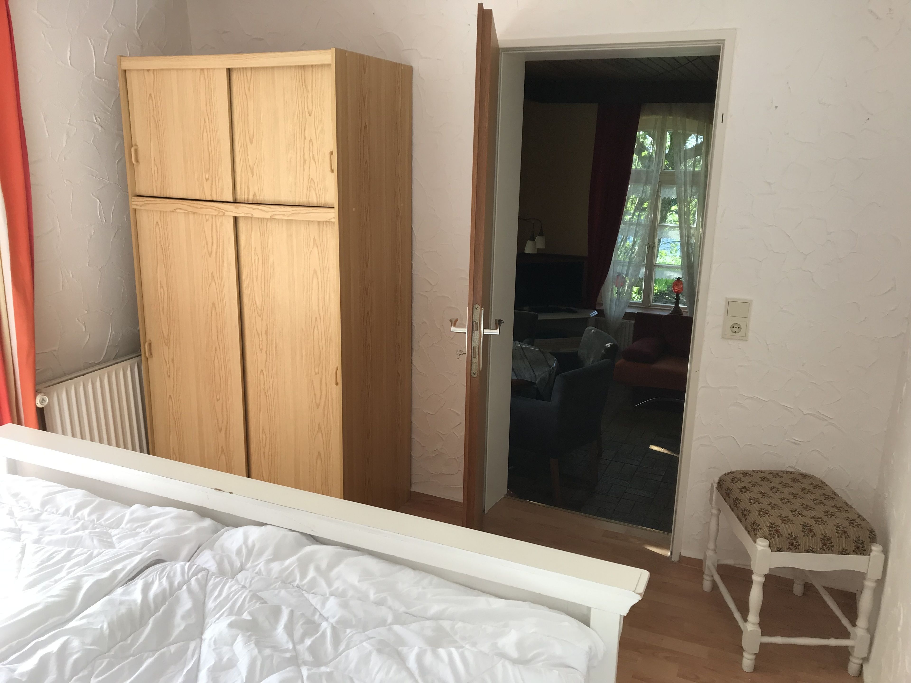 Un dormitorio con una cama, un armario de madera, un taburete pequeño y una puerta que da a una sala de estar.