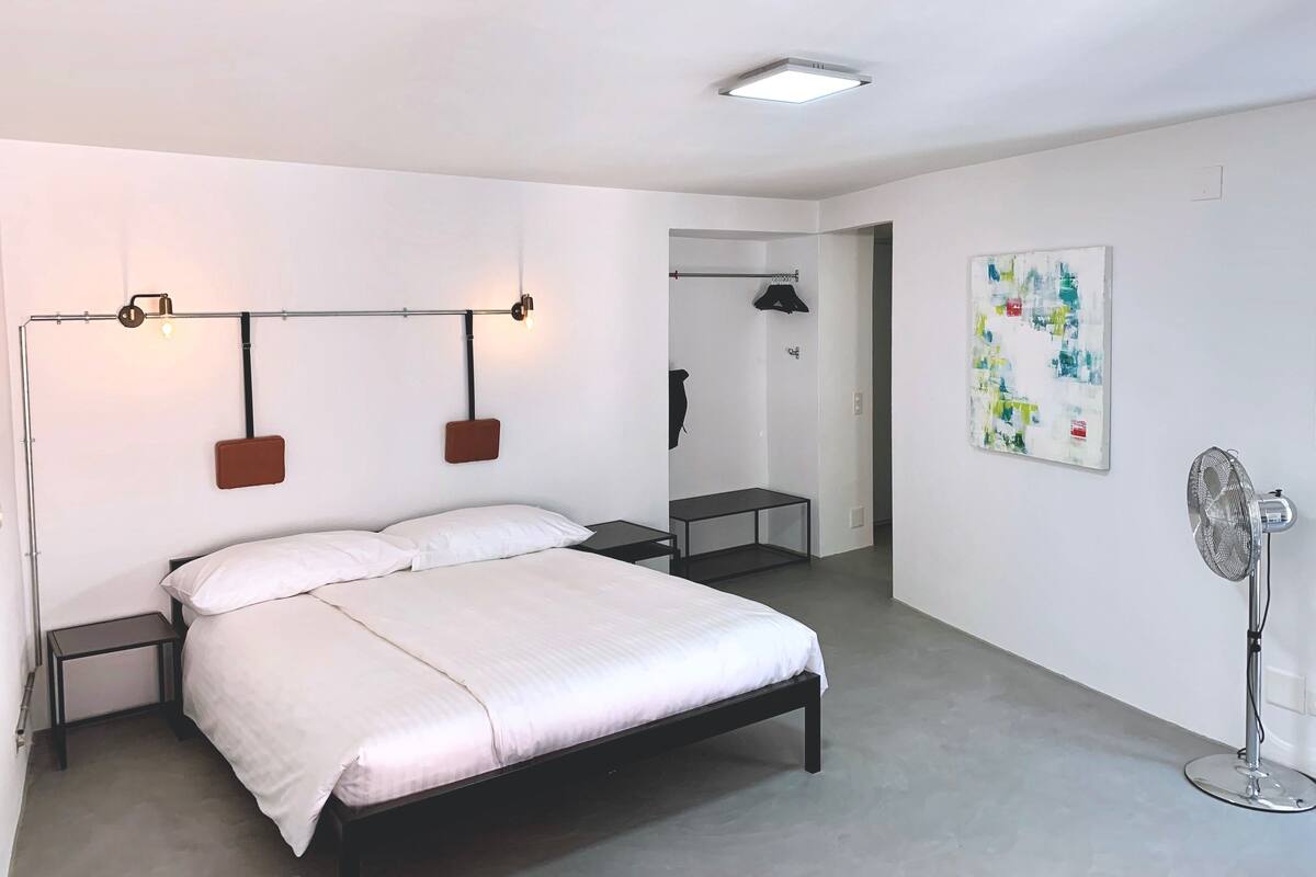 A modern bedroom with a bed, bedside tables, a hanging light fixture, a ceiling light, a wall-mounted fan, and a door leading to another room.
