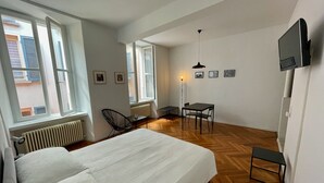 A bedroom with a large bed, a desk, and a chair. There are windows and a radiator.